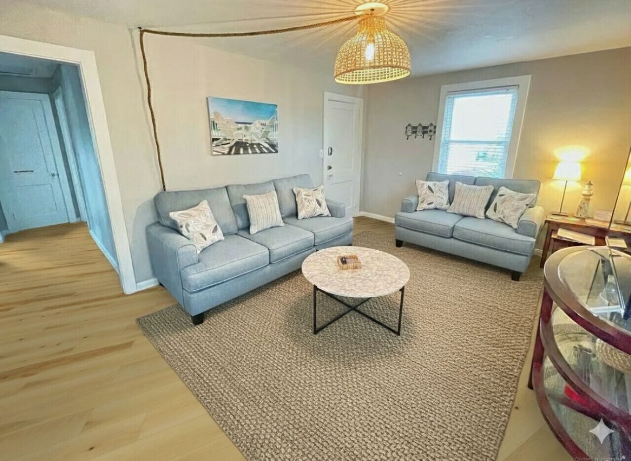 Un salon avec deux canapés bleu clair, une table basse ronde, un tapis à motifs, un ventilateur de plafond et un tableau accroché au mur.