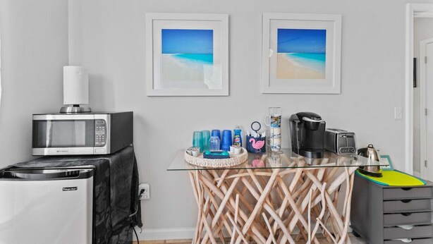 A kitchenette with a microwave, toaster, and coffee maker, a glass table with a woven base, and two framed beach pictures on the wall.