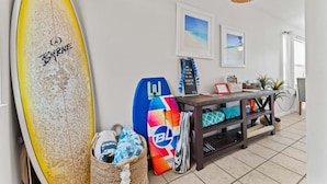 A room with a surfboard leaning against the wall, a colorful surfboard on a shelf, and a beach-themed sign.