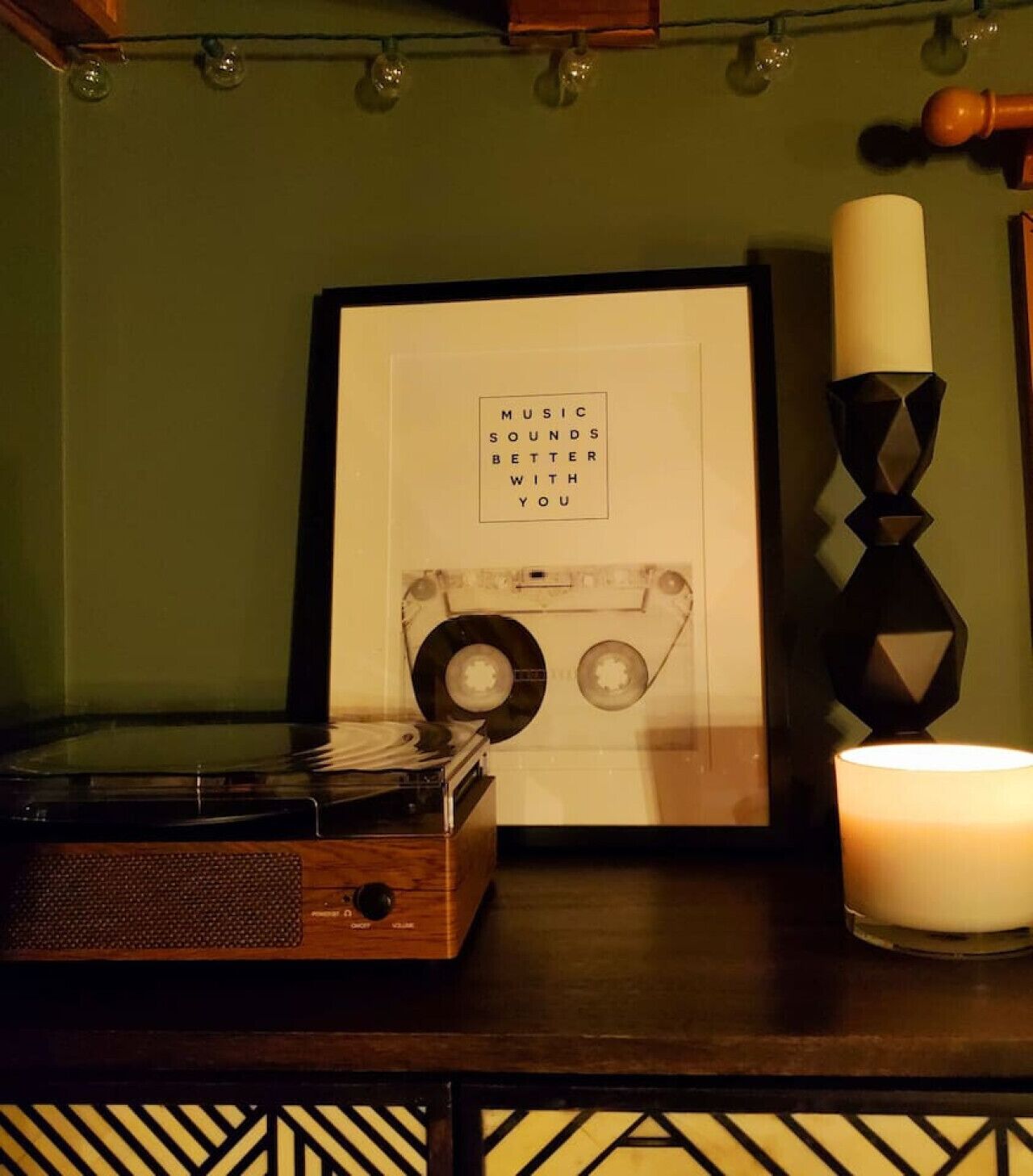 En vintage pladespiller, et indrammet billede med teksten ”MUSIC SOUNDS BETTER WITH YOU”, og et lys på et bord.