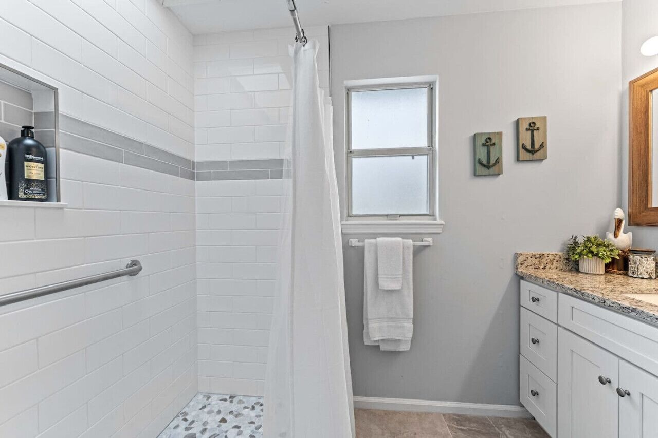 Une salle de bain avec des murs recouverts de carreaux blancs, une douche avec un rideau blanc, une fenêtre et un meuble-lavabo avec un plan de travail.
