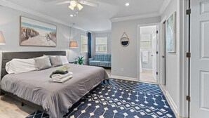 A bedroom with a bed, a ceiling fan, a painting, a blue sofa, and a patterned rug. - Steps 2 Sand! Bch amenities, yard, rooftop & games (Jacksonville Beach)