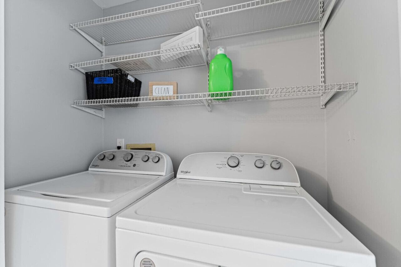 A laundry area with two white Whirlpool washers, metal shelves above holding baskets and a green bottle, and a 'CLEAN' sign.