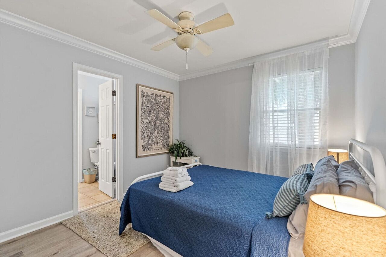 A bedroom with a bed, a ceiling fan, a window with sheer curtains, a bedside lamp, and a framed artwork on the wall.