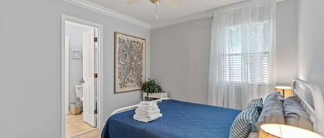 A bedroom with a bed, a ceiling fan, a window with sheer curtains, a bedside lamp, and a framed artwork on the wall.