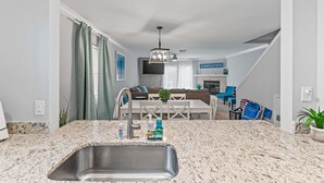 A modern kitchen with granite countertops, a stainless steel sink, and a dining area with a white table and blue chairs. - Steps 2 Sand! Bch amenities, yard, rooftop & games (Jacksonville Beach)