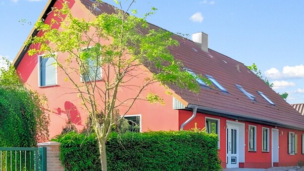 Ein rotes Haus mit geneigtem Dach, einem Baum davor und einer Hecke.