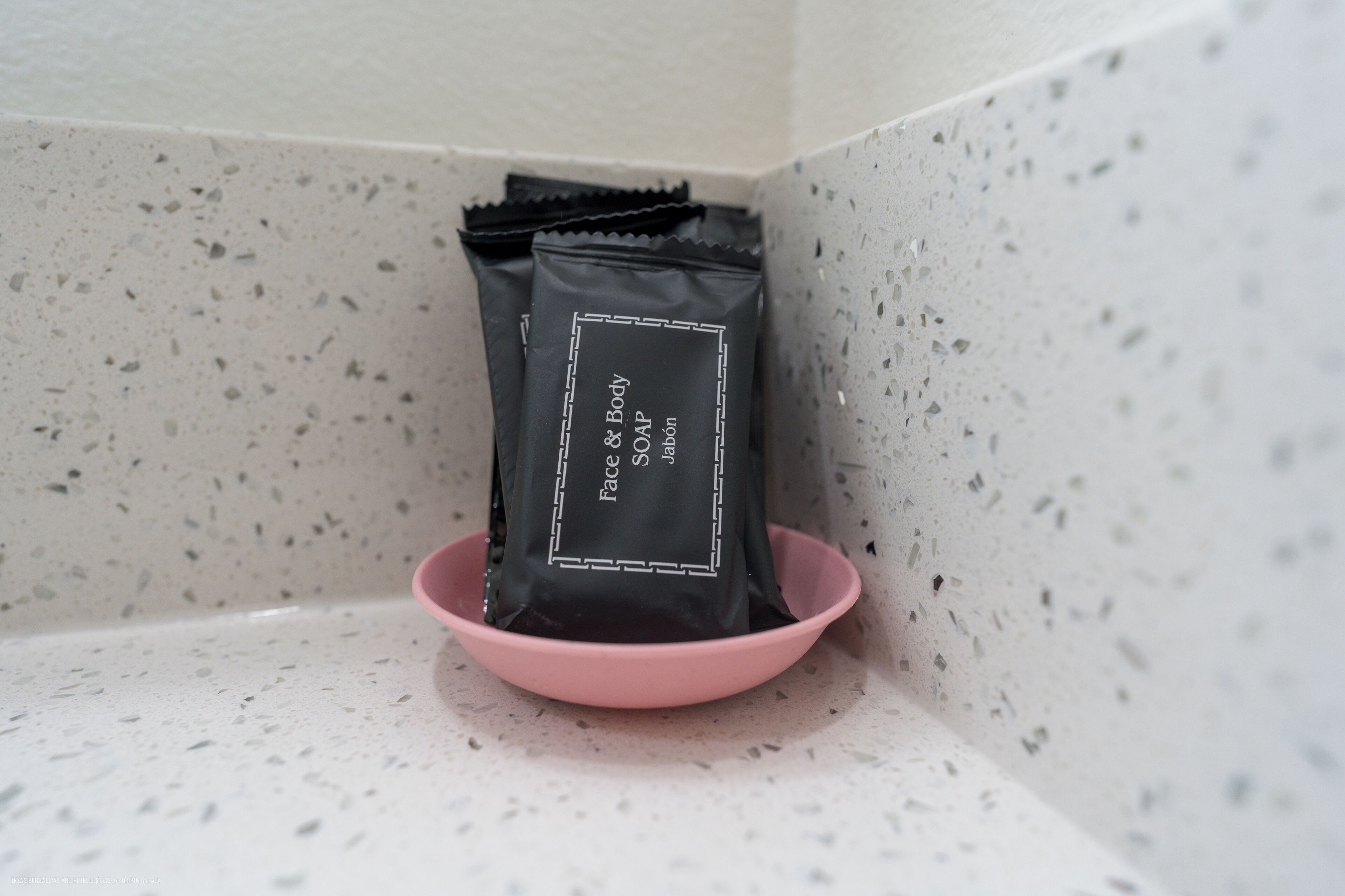 Un coin de pièce où se trouve un bol rose contenant des sachets noirs étiquetés « Savon pour le visage et le corps ».