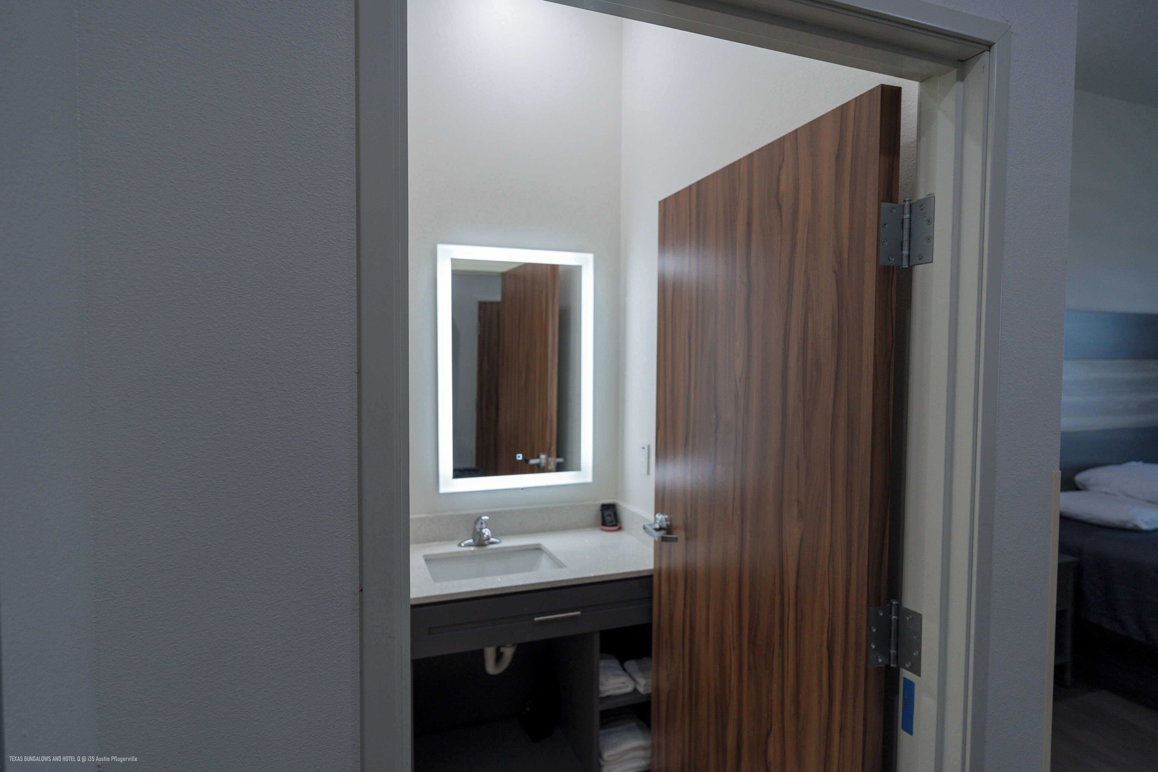 A modern bathroom with a wooden door, a white sink, and a mirror with a lighted frame.