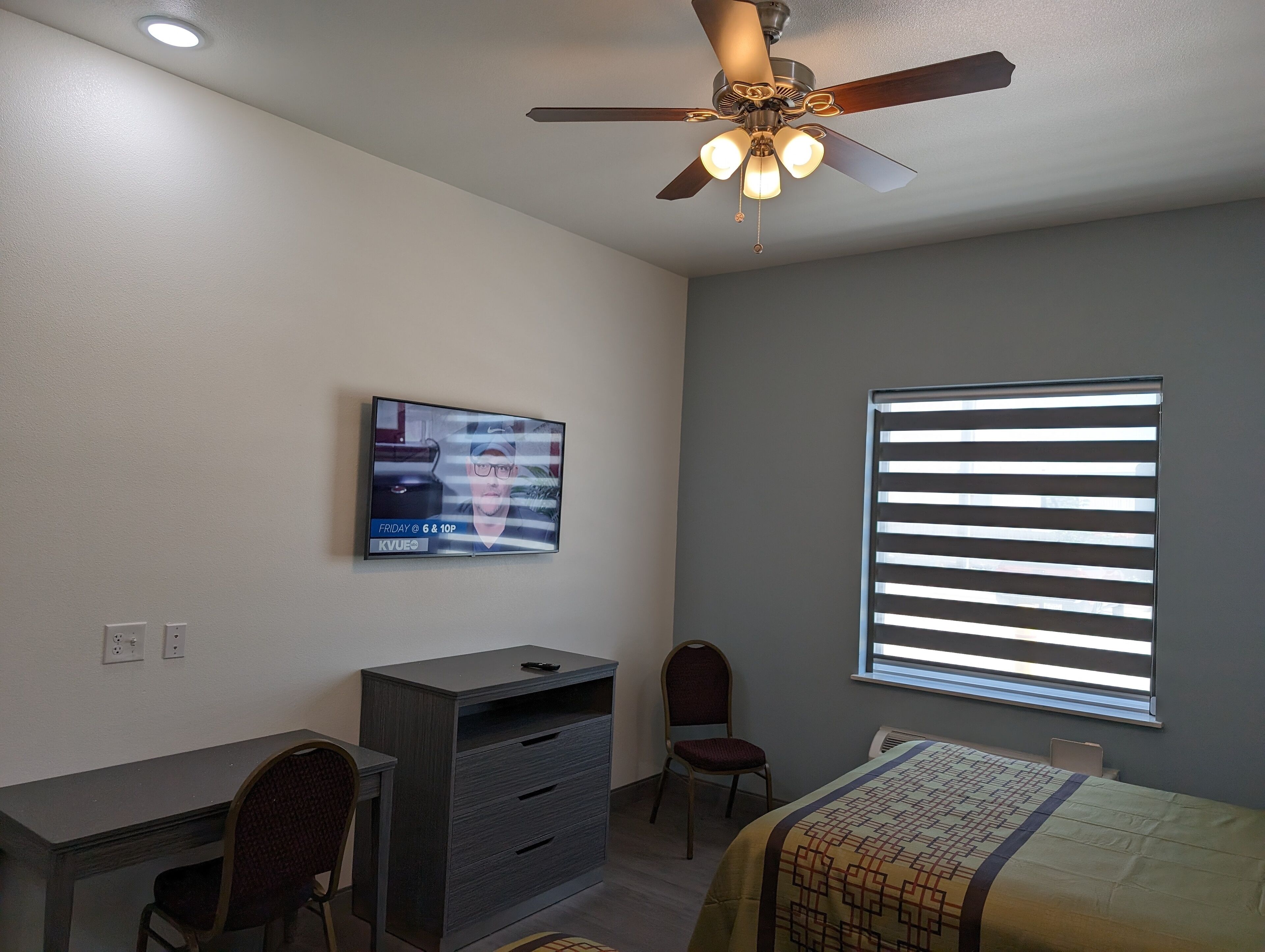 Quarto com escrivaninha, cadeira, cama, ventoinha no teto, televisão e janela com persianas.