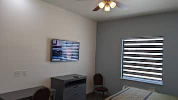 Quarto com escrivaninha, cadeira, cama, ventoinha no teto, televisão e janela com persianas.