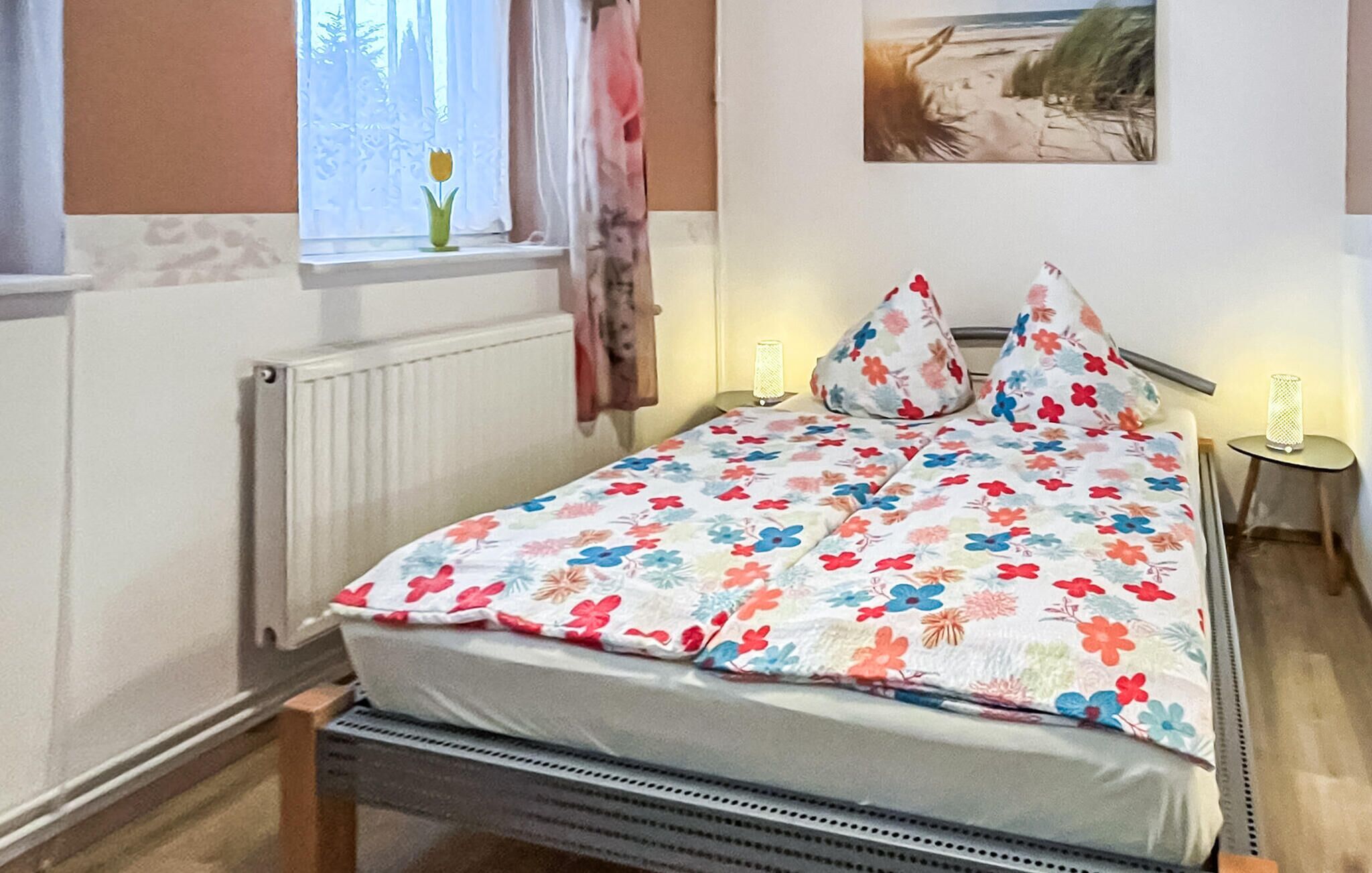 Un dormitorio con colcha estampada, un radiador, una ventana con cortina, una mesita de noche con lámpara y un cuadro en la pared.