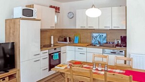 A compact kitchen with white cabinets, a microwave, a clock, and a dining area with a wooden table and chairs. - Ferienwohnung Dierhagen (Dierhagen (Ostseebad))