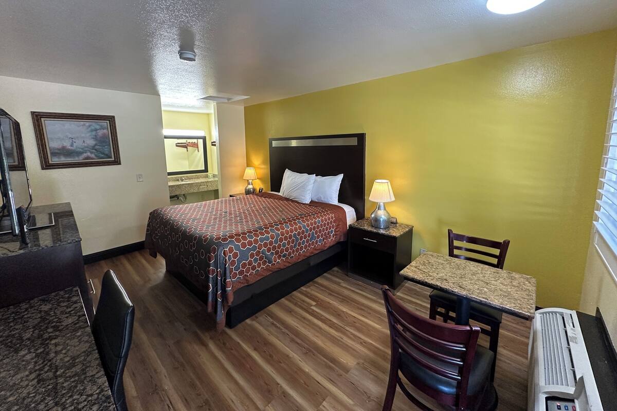 A hotel room with a bed, bedside table with lamp, wooden floor, and a desk with chair.