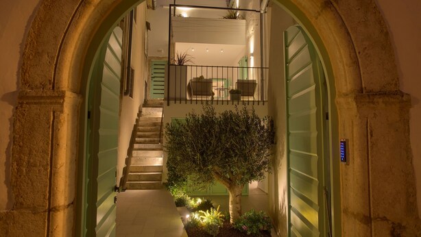 Un couloir étroit surmonté d’une arche en pierre, un escalier menant à un balcon et un petit arbre au centre.