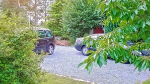 A gravel driveway leading to a parked car, surrounded by lush greenery and a red door.