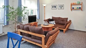 A living room with a brown sofa, a blue chair, a coffee table, and a TV. There are two framed pictures on the wall and a shelf with cups.
