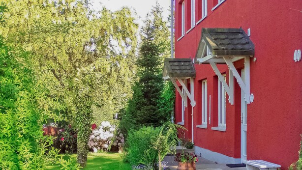 A red building with white trim, a covered porch, and a garden with various plants and flowers.