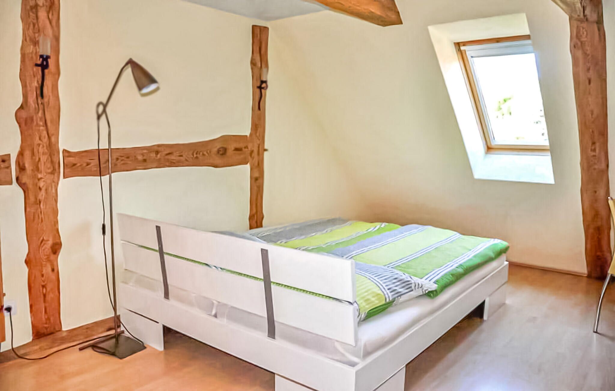A small, single-bed room with a sloped ceiling, exposed wooden beams, a window, and a bedside lamp.