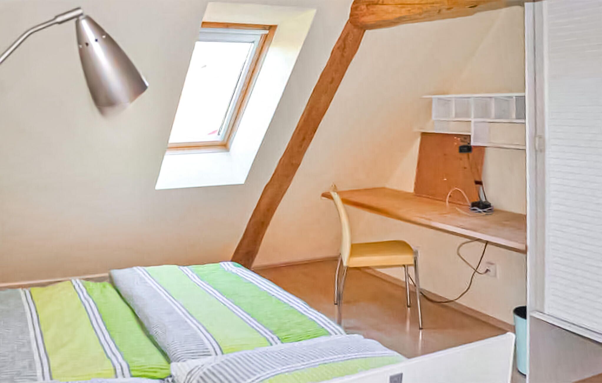 A small, well-lit room with a sloped ceiling, a single bed with striped bedding, a desk with a chair, a window, and a desk lamp.
