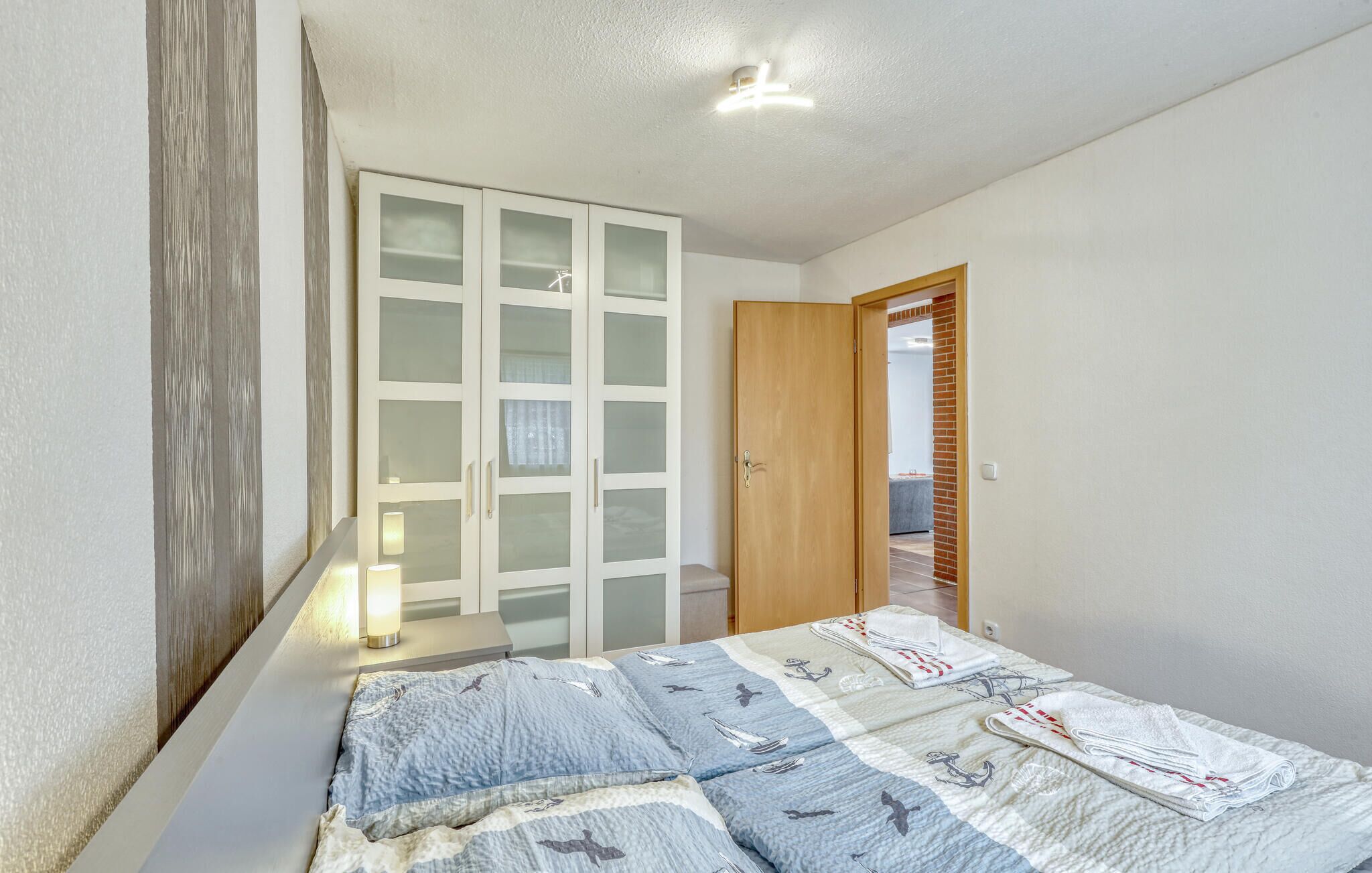 Un dormitorio con cama, armario de puertas correderas con vidrio esmerilado, una puerta de madera y una mesita de noche con lámpara.