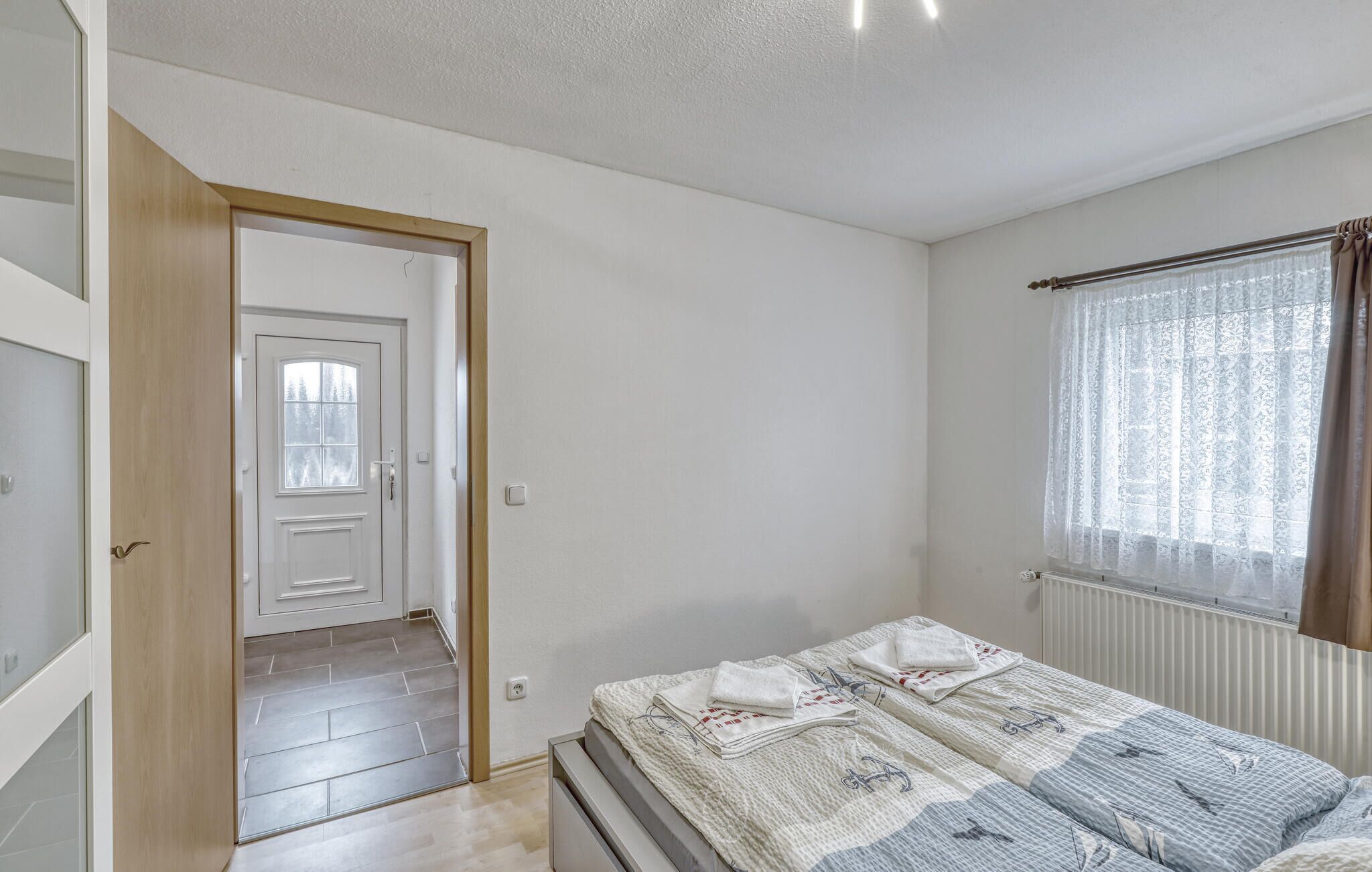 Een slaapkamer met een bed, een raam met gordijnen, een radiator en een deur naar een andere kamer.