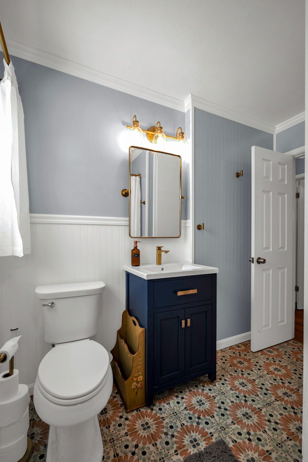 A bathroom with a white toilet, a dark blue vanity with gold handles, a mirror with lights, and patterned floor tiles.