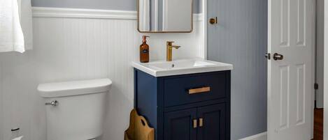 A bathroom with a white toilet, a dark blue vanity with gold handles, a mirror with lights, and patterned floor tiles.