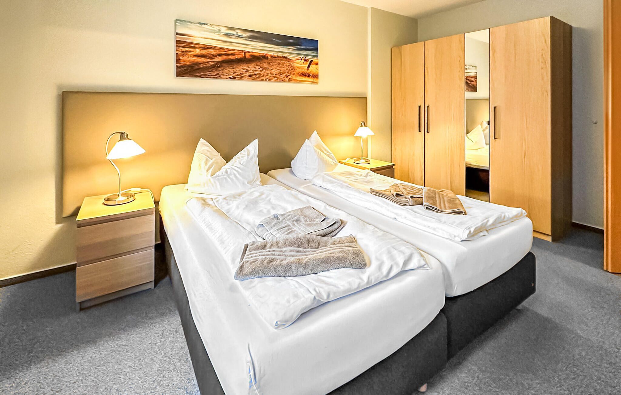 A double bed with white linens, two bedside tables with lamps, a wooden wardrobe, and a beach-themed painting on the wall.