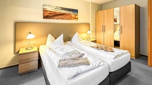 Un lit double avec du linge de lit blanc, deux tables de chevet avec des lampes, une armoire en bois et un tableau sur le thème de la plage accroché au mur.