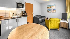 Une cuisine compacte équipée d’un four à micro-ondes, d’une machine à café et d’un évier. On y trouve une table ronde en bois, un canapé gris et un fauteuil jaune. Un téléviseur à écran plat est posé sur un meuble en bois.