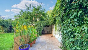 En smal stentrappe fører til et overdækket udendørs siddeområde med bord og stole, omgivet af frodig vegetation og krukkeplanter.