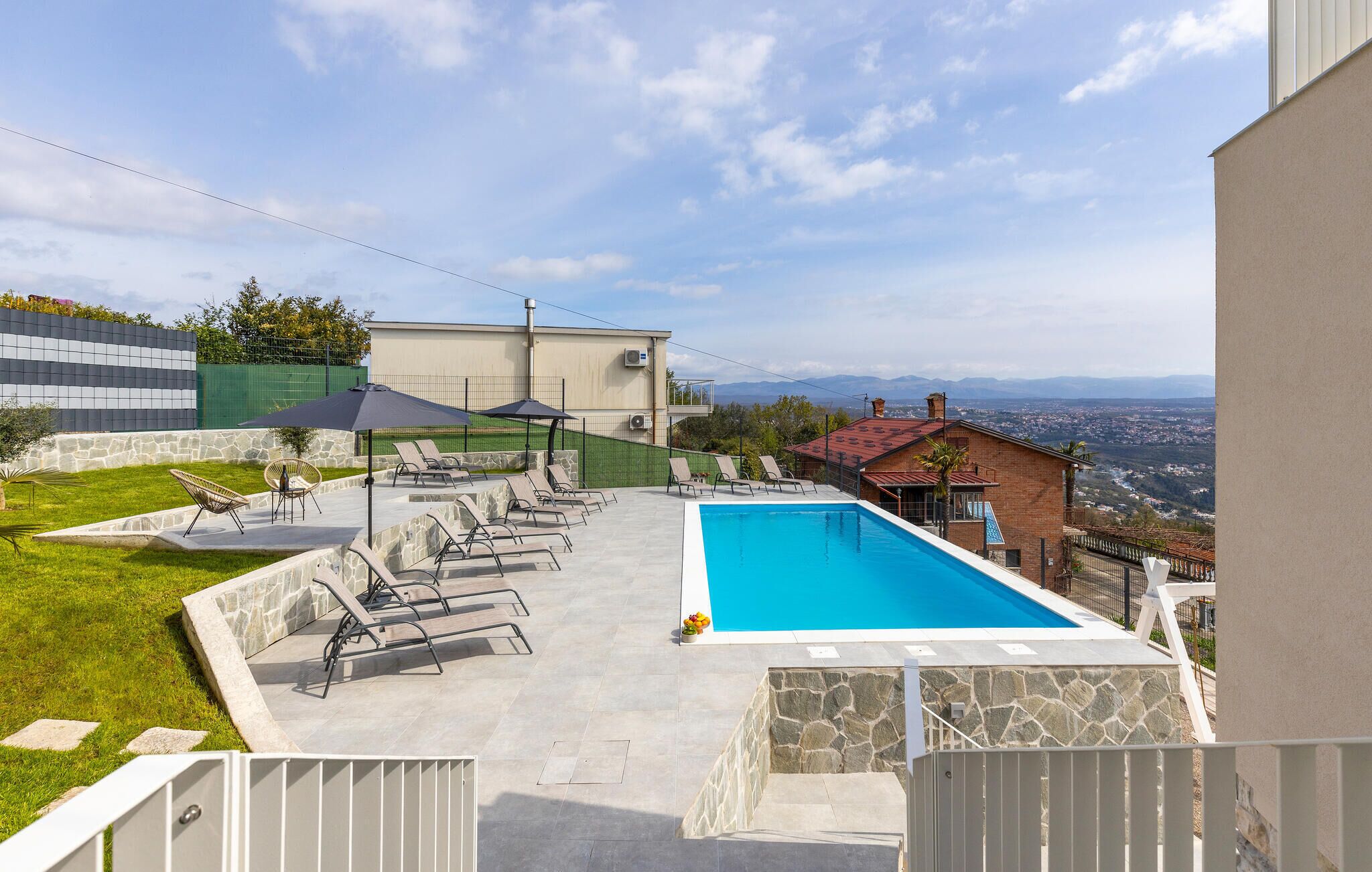 A swimming pool with lounge chairs and umbrellas, surrounded by stone walls and a view of a cityscape.