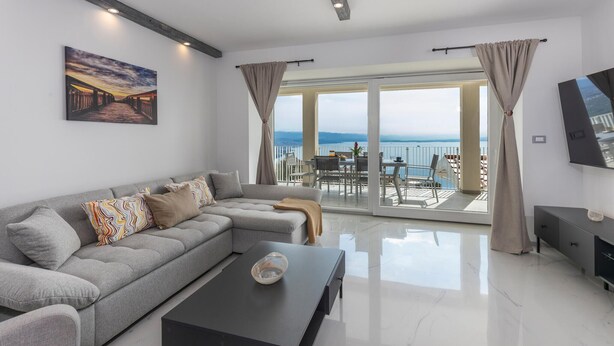 Ein modernes Wohnzimmer mit grauem Sofa, einem Couchtisch und einem Gemälde an der Wand. Ein Balkon mit Tisch und Stühlen bietet Blick auf das Meer.