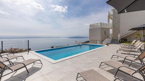 Ein Pool auf dem Dach mit Blick auf das Meer, umgeben von Liegestühlen und einem modernen Gebäude mit Steinaußenwand.