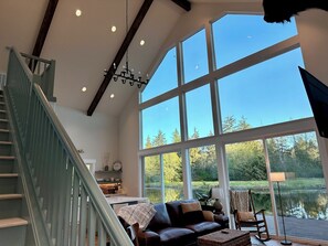 A spacious living room with a large window overlooking a lake, a staircase, and a cozy seating area. - Gorgeous cabin, secluded fishing pond, swimming, paddling. Central for Daytrips! (Elma)