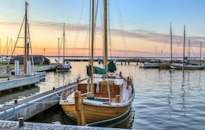 Um barco de madeira atracado em um porto, com outros barcos, um cais e um pôr do sol ao fundo.