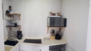 Een kleine keuken met een magnetron, een koffiezetapparaat en een waterkoker op het aanrecht. Er staan planken met servies en potten aan de muur.
