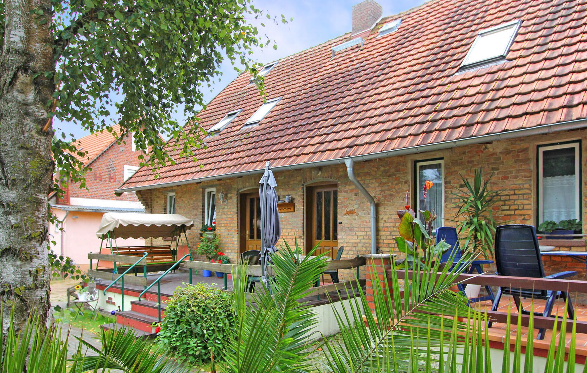 Ein Ziegelhaus mit rotem Ziegeldach, einer kleinen Veranda mit Gartenmöbeln und einem Baum im Vordergrund.