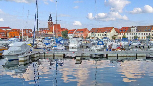 Ein Yachthafen mit vertäuten Booten, ein markanter Turm im Hintergrund und bunte Gebäude entlang der Uferlinie.