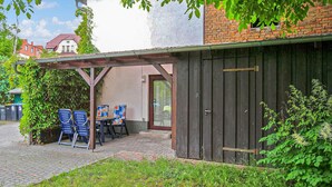 A covered outdoor seating area with blue chairs and a table, a wooden door, and a brick building in the background. - Cozy apartment in Waren (Müritz) (Waren (Müritz))