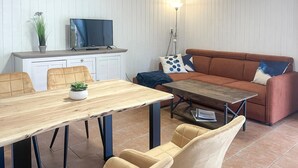 A living room with a wooden dining table, beige chairs, a brown sofa with patterned cushions, a television on a white cabinet, and a floor lamp.