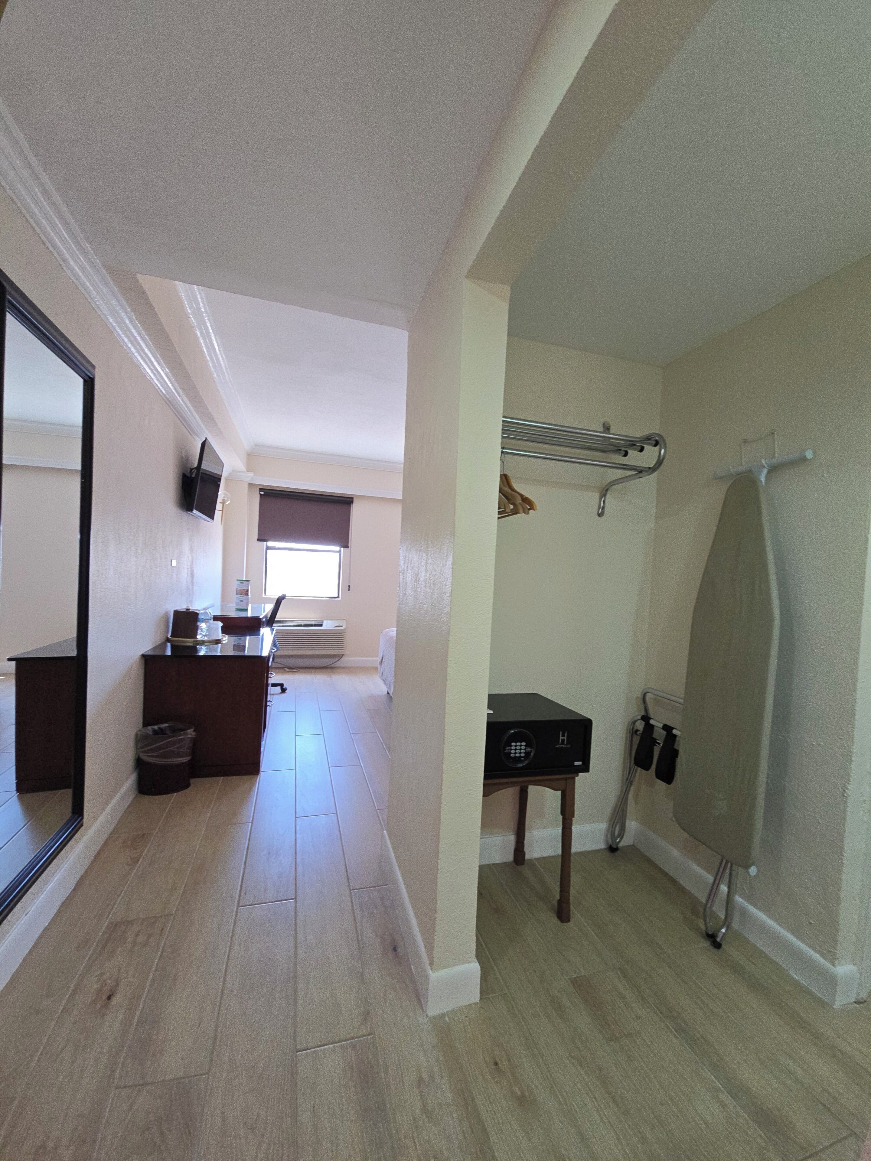 Quarto de hotel com piso de madeira, uma escrivaninha com cadeira, TV fixada na parede, área de chuveiro com porta-toalhas e uma pequena mesa com rádio.
