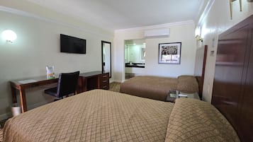 Habitación de hotel con dos camas, un escritorio, una silla, un televisor y un baño visible a través de una puerta abierta.