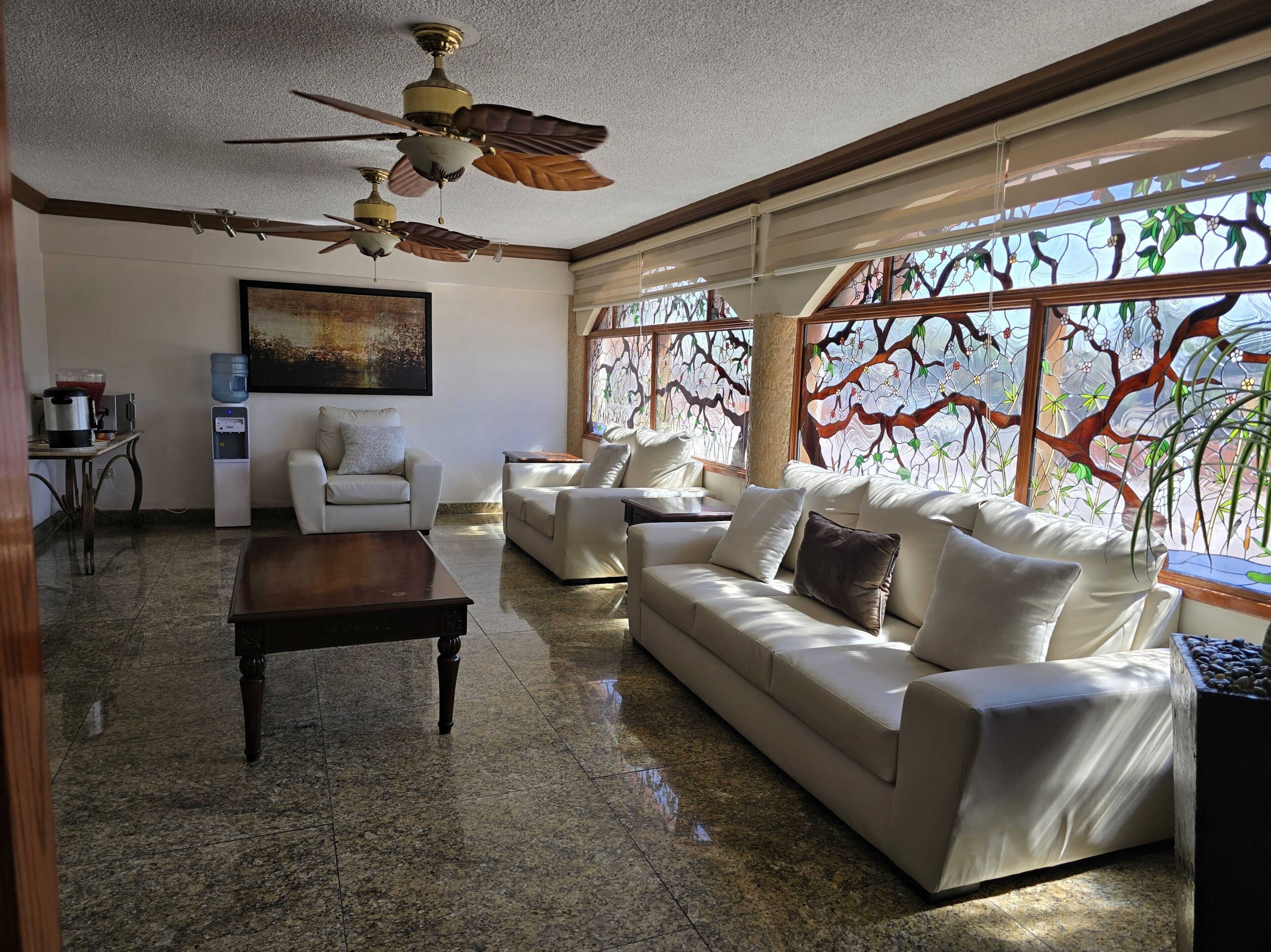 A spacious living room with a large sofa, a coffee table, and a ceiling fan. There is a painting on the wall and a window with a tree view.