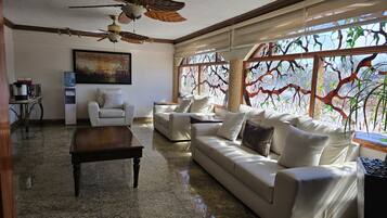 A spacious living room with a large sofa, a coffee table, and a ceiling fan. There is a painting on the wall and a window with a tree view.