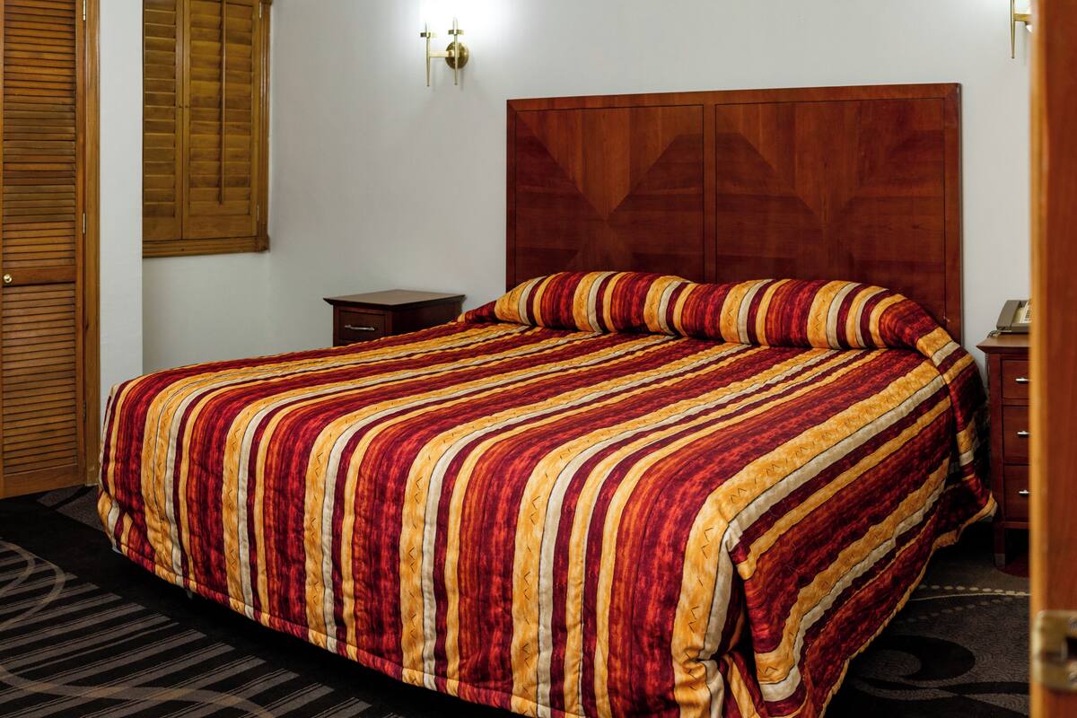 A neatly made bed with a striped bedspread, a wooden headboard, a nightstand with a lamp, and a door with a wooden shutter.
