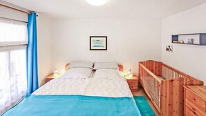 Ein Schlafzimmer mit einem Bett, einem Babybett, einem Nachttisch, einer Wandregal und einem Fenster mit Vorhängen.