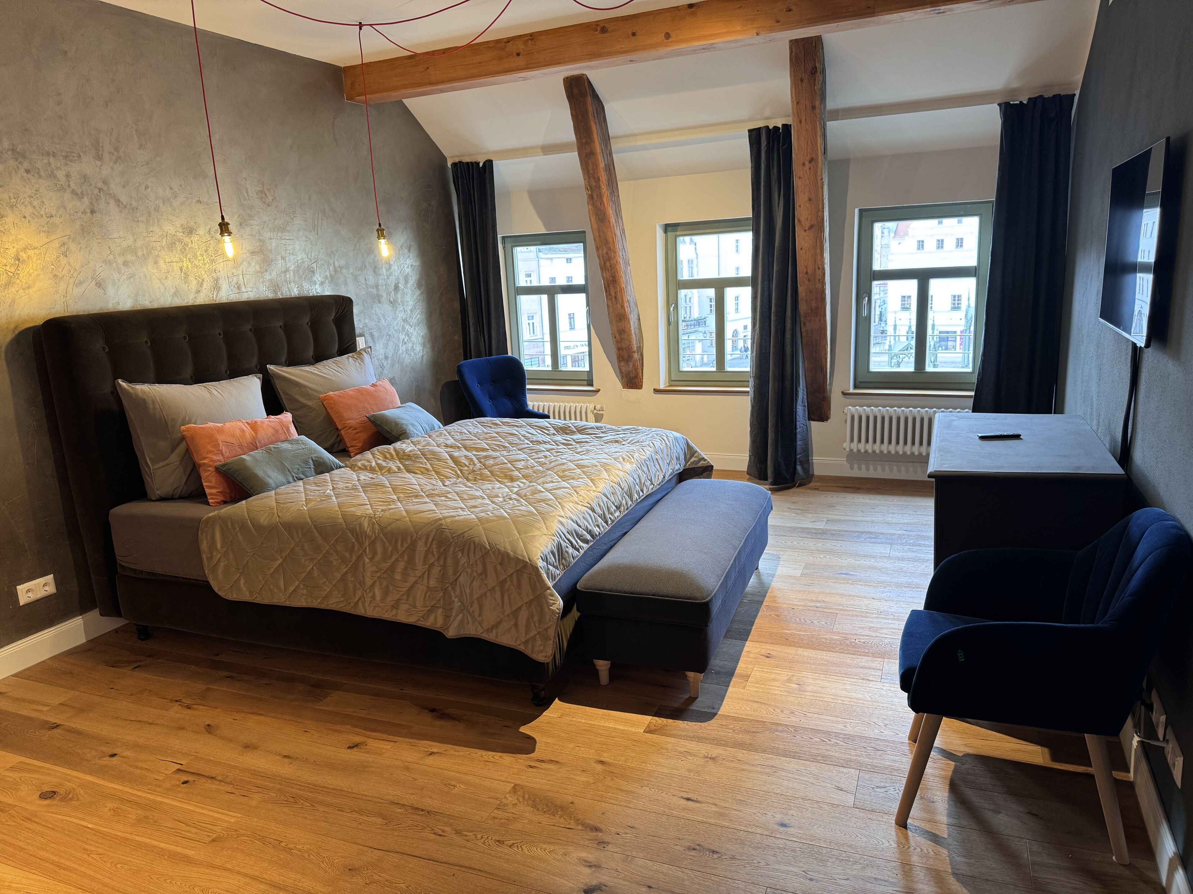 A bedroom with a large bed, a desk, and a chair. There are windows with curtains, a radiator, and a TV on the wall.
