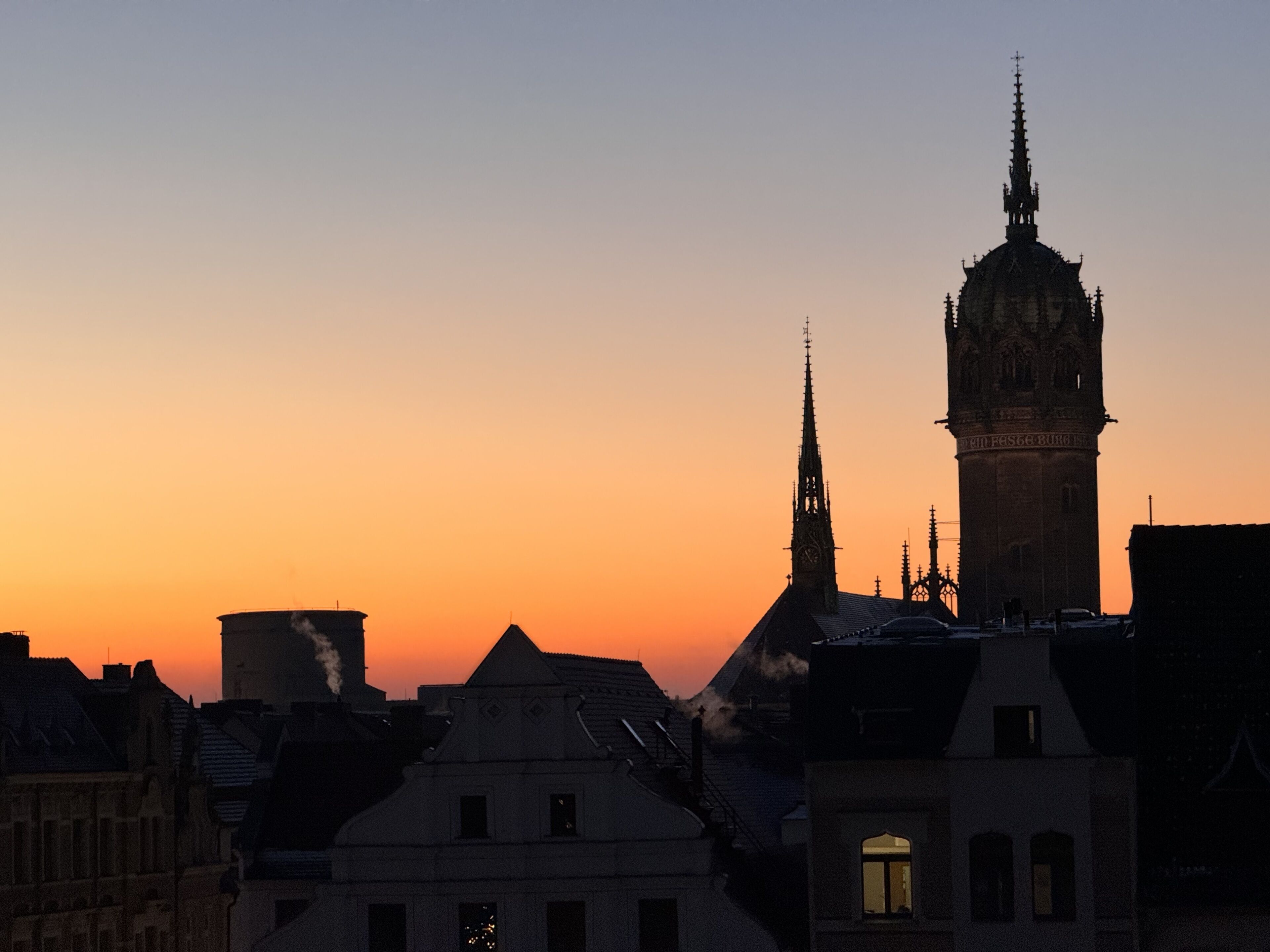 Eine Stadtansicht im Abendlicht mit markanter historischer Architektur, darunter ein hoher Kirchturm und ein Glockenturm.